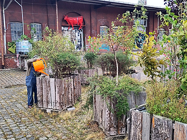 Auf einem Platz stehen grob gezimmerte Pflanzenkästen, eine Person gießt die Pflanzen mit einer orangenen Gießkanne