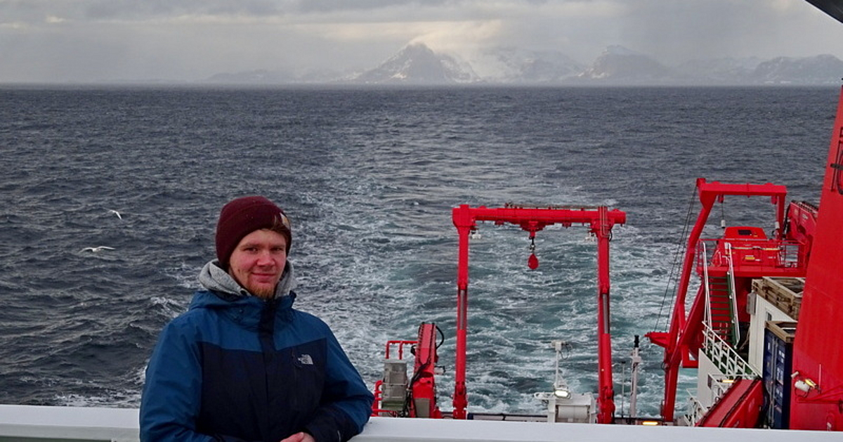 Ein Wissenschaftler steht mit blau-schwarzer Jacke und Wollmütze an Deck des Forschungsschiffs Maria S. Merian. Im Hintergrund ist das Heck des Schiffes und die Südspitze Grönlands zu sehen.