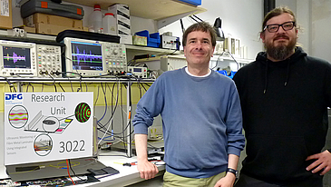 Two people stand in front of a desk in a laboratory and look into the camera. There is a lot of technical equipment on the desk and on the wall behind them. Some cables can be seen. A graphic next to them reads "Research Uni 3022.