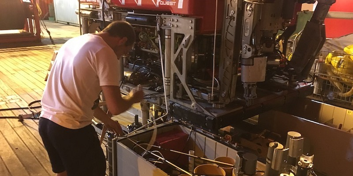 A researcher works on the water samplers attached to the red, remotely operated underwater vehicle MARUM Quest 4000, which sits on the research vessel's brightly lit working deck.