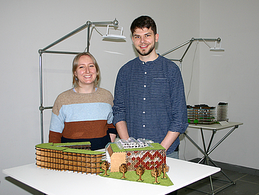 Junge Frau und junger Mann stehen vor einem Tisch mit einem 3D-Modell von einem Gebäude.