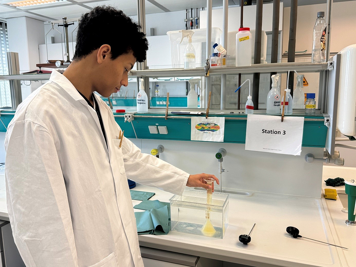 Ein Schüler in Kittel steht an einer Laborbank betrachtet einen Versuch zur CO2-Löslichkeit in Wasser.