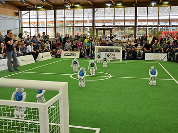 Roboter spielen Fußball, Publikum schaut zu.