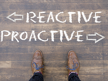 Man looking down at the words "REACTIVE" and "PROACTIVE" written on the floor Ein Mann schaut auf die Wörter "Reactive" und "Proactive" herab, die mit Kreide auf den Boden geschrieben wurden