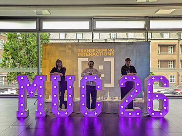 Lisa, Kathi, and Rachel are posing behind large letters that spell out “MUC 26.”