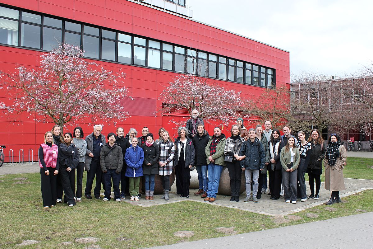 Grupppenbild von Teilnehmer:innen eines internationalen Symposium an der Universität Bremen stehen vor dem MZH, im Hintergrund Kirschbäume in beginnender Blüte