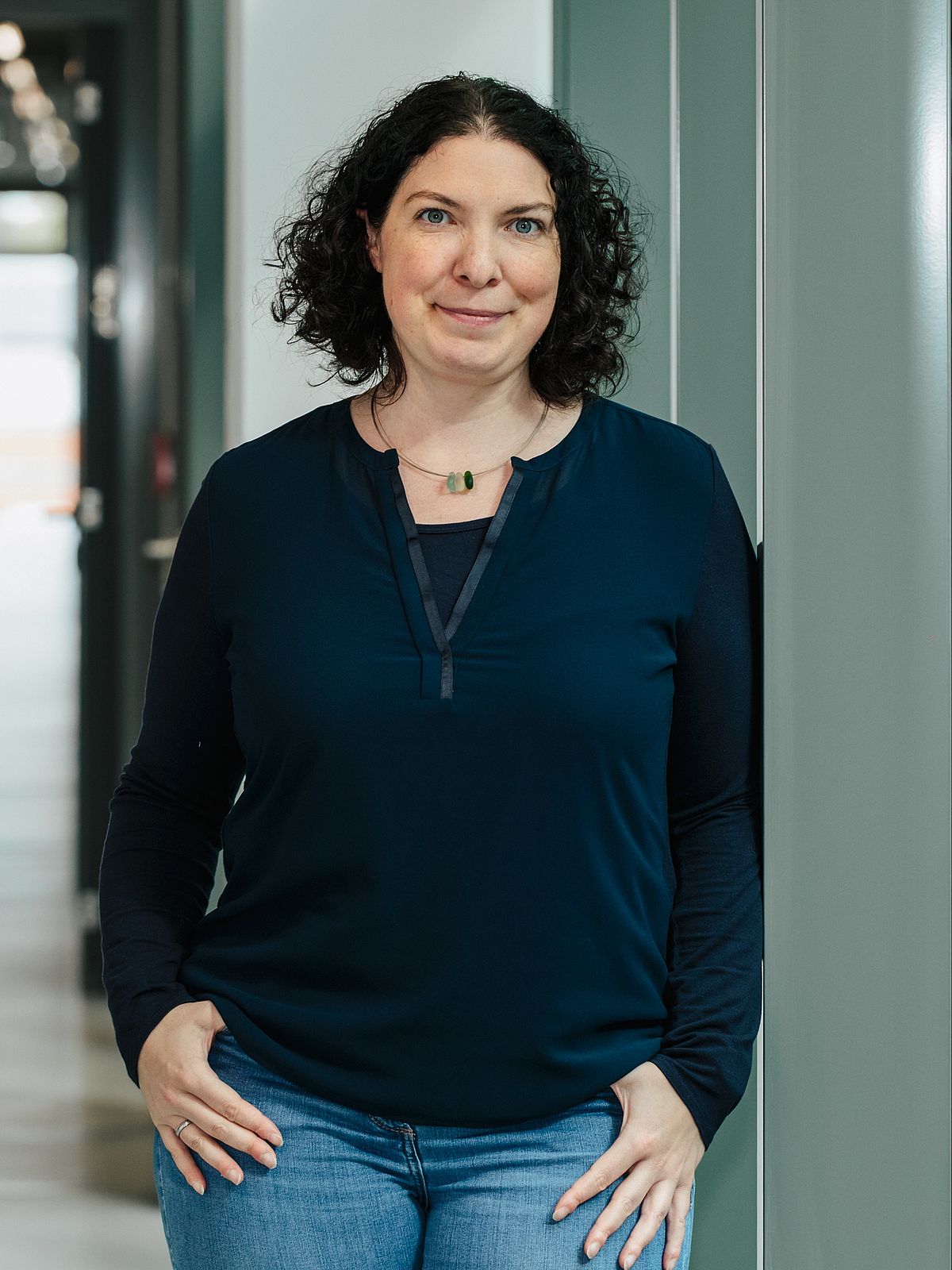 Christina Roggatz steht auf einem Flur und schaut freundlich in die Kamera. Sie ist von der Taille bis zum Kopf mit den lockigen dunklen Haaren zu sehen.