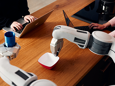 Ein Roboter hält eine Schüssel fest, die auf einem Tisch steht. Im anderen Arm hält er einen Becher, aus dem er offensichtlich etwas ausschütten will.