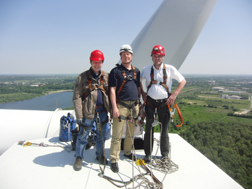 Drei Männer stehen auf der Gondel einer Windenergieanlage. Sie tragen Klettergurte und Schutzhelme.