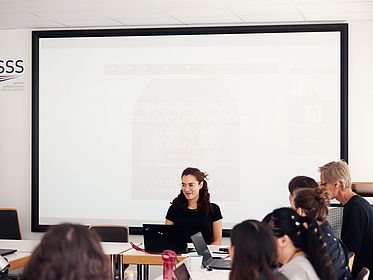Eine Frau hält einen Vortrag, am rechten Bildrand sind weitere Personen zu sehen, die dem Vortrag zuhören.