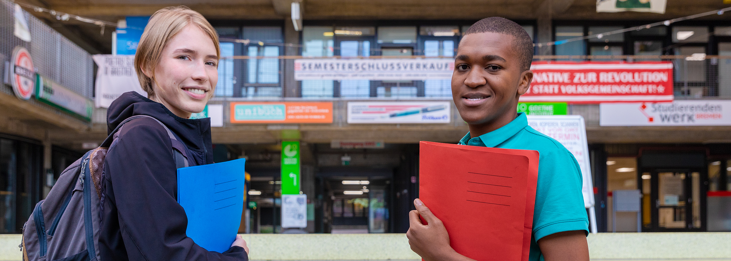 Zwei Studierende stehen mit Mappen in der Hand in der Glashalle