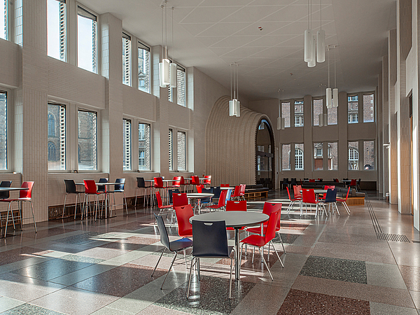 In a large hall with wood paneling on the wall, there are a few tables with blue and red chairs around them
