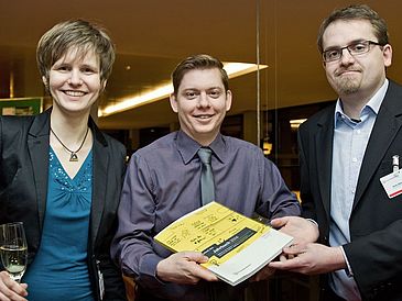 Zwei Männer und eine Frau halten ein gelbes Buch in die Kamera.