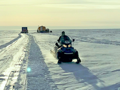 A scientist rides a snowmobile through a landscape of snow and ice. There are buildings in the background.