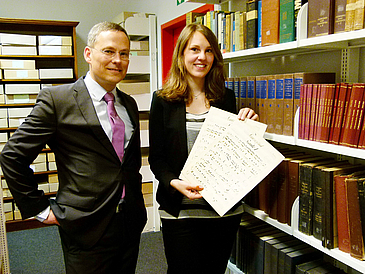 Mann und Frau mit Notenblättern vor Bücherregalen.