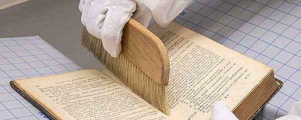 A gloved hand runs a brush through the crease of a book.