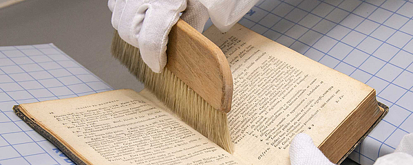 A gloved hand runs a brush through the crease of a book.