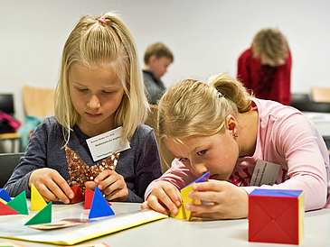 Zwei Mädchen sitzen an einem Tisch und arbeiten mit bunten geometrischen Formen. Im Hintergrund sieht man weitere Kinder.