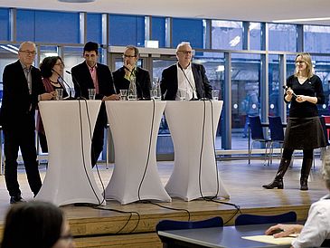 Fünf Personen stehen an Tischen, rechts die Moderatorin