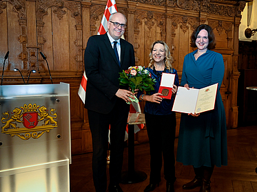 Drei Personen stehen in einem prunkvollen Raum mit geschnitzten Holzwänden. Links steht ein großer, glatzköpfiger Mann mit Brille in einem dunklen Anzug, der einen Blumenstrauß überreicht. In der Mitte eine kleine Frau mit blonden Haaren, die eine Medaille hält und lächelt. Rechts eine Frau mit schulterlangen dunklen Haaren in einem blauen Kleid, die eine Urkunde präsentiert. Im Vordergrund ist ein Rednerpult mit dem Wappen von Bremen, links im Hintergrund eine rot-weiß-karierte Fahne.