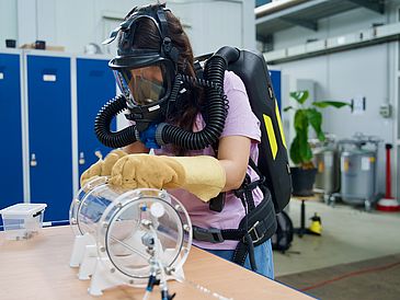 Eine Frau mit Sauerstoffgerät, Atemmaske und dicken Handschuhen montiert ein Bauteil.