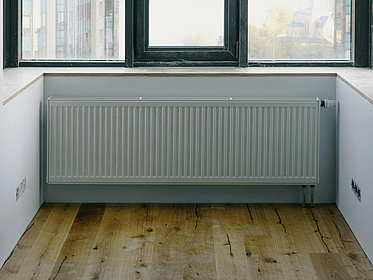 Ein weißer Heizkörper hängt in einer Wohnungsnische unter einem Fenster an der Wand