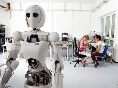 Ein humanoider Roboter in einem Labor mit zwei Personen im Hintergrund.