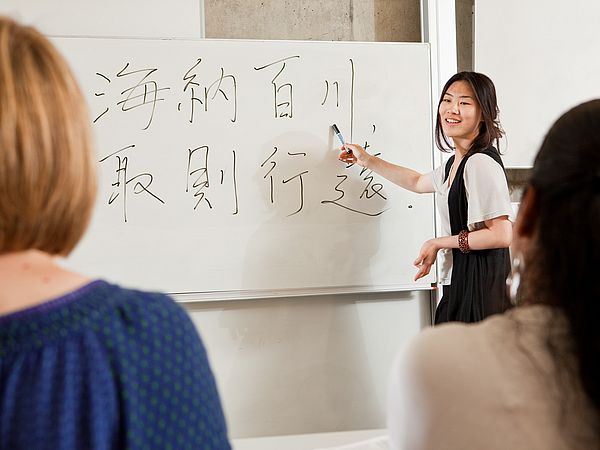 Eine Lehrende steht vor einer Tafel mit Text in mehreren Sprachen.