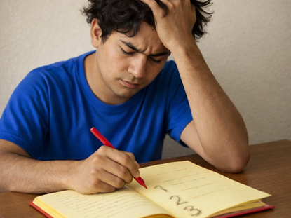 Eine Person sitzt an einem Tisch vor einem aufgeschlagenen Buch, das auf braunem Untergrund liegt. Die Person trägt ein blaues T-Shirt und hält einen Stift in der rechten Hand, mit dem sie auf die Buchseite schreibt. Die linke Hand ist an den Kopf gelegt und die Person rauft sich die Haare als Zeichen von Nachdenklichkeit oder Verzweiflung.