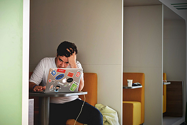 Das Foto zeigt einen jungen Mann, der an seinem Laptop sitzt in einer Sitz- und Arbeitsecke eines größeren Gebäudes und sich dabei die Haare rauft.