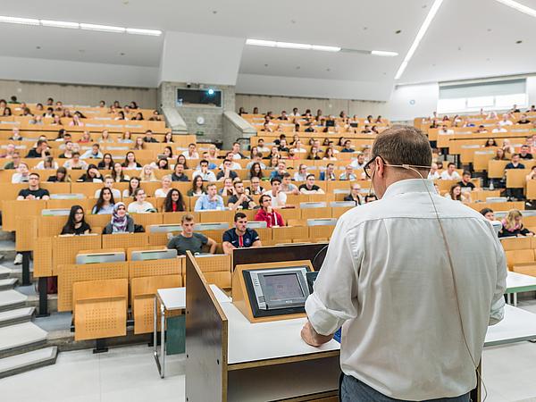 A lecturer speaks to an audience in a packed lecture hall.