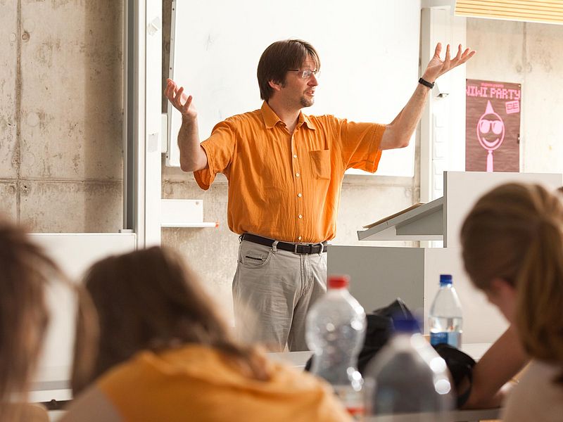 A teacher speaks in front of an audience in a seminar room.