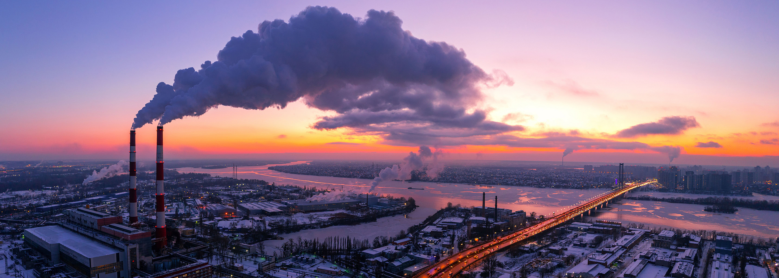 Luftbild einer Stadt am Fluss bei Sonnenaufgang. Aus hohen Fabrikschornsteinen steigt eine große Menge Rauch auf.