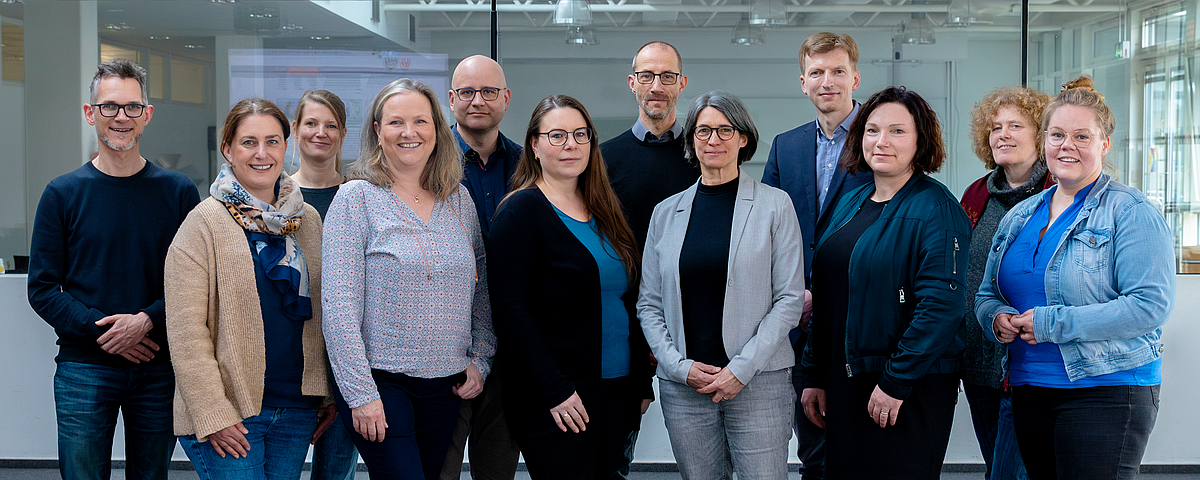 Gruppenfoto des Referat 13 der Universität Bremen. Zwölf Personen, in zwei Reihen hintereinander versetzt aufgestellt, schauen zur Betrachterin / zum Betrachter.