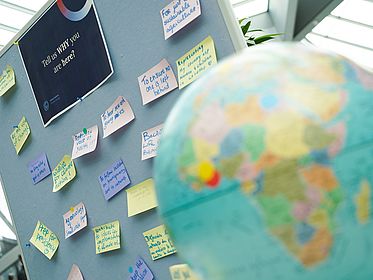 Ein Globus steht unscharf im Vordergrund. Dahinter hängt eine Pinnwand mit der Frage „Tell us why you are here?“ und mehreren handschriftlichen Notizzetteln zu Klima, Gesundheit und Nachhaltigkeit