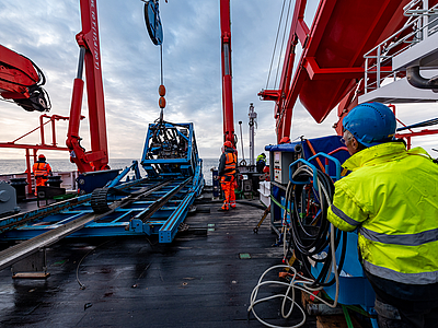Mehrere Menschen stehen auf einem Schiffsdeck und lassen mithilfe eines Krans ein Meeresboden-Bohrgerät zu Wasser.