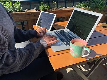 Eine Person arbeitet auf dem Balkon an ihrem Laptop und trinkt Kaffee.