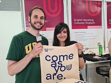Zwei Studierende stehen lächelnd im Vordergrund und halten eine Tasche mit der Aufschrift "Come as you are" hoch. Im Hintergrund sieht man Plakate des Standes des Fachbereichs 10 auf der Informationsveranstaltung für SchülerInnen.
