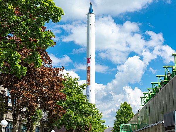 Ein Banner hängt am Fallturm, das auf die Ausstellung "WARUM? DARUM!" anlässlich des 50-jährigen Bestehens der Universität hinweist.