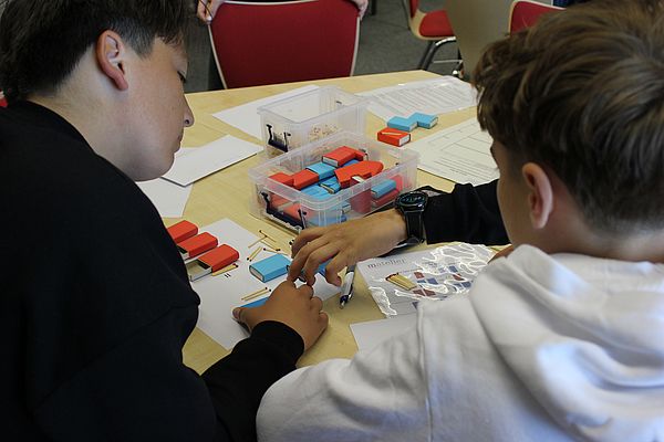 Zwei Schüler sind von hinten zu sehen, außerdem ragt die Hand einer weiteren Person ins Bild. Auf dem Tisch vor den Schüler:innen liegen Streichholzschachteln und Streichhölzer.