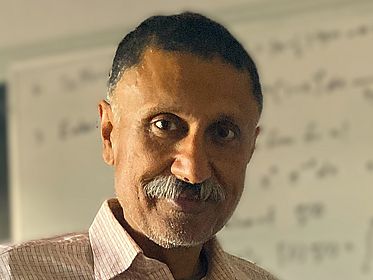 Bild von Bharath Sriraman. Er schaut freundlich in die Kamera, im Hintergrund leicht unscharf ein mit mathematischen Symbolen beschriebenes Whiteboard