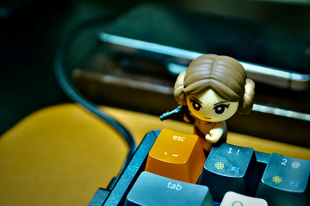 Eine kleine Plastikspielzeugfigur steht hinter einer Tastatur. Die Escape-Taste sticht durch ihre gelbe statt graue Farbe hervor. Dahinter nimmt die Figur Deckung.