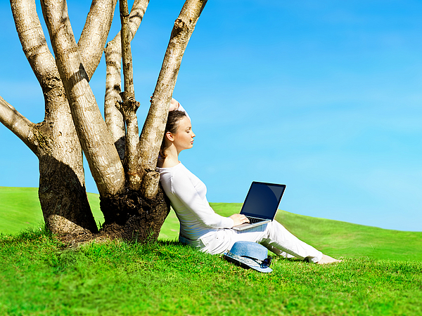Frau, die mit einem Laptop auf dem Schoß an einen Baum gelehnt auf der grünen Wiese sitzt.