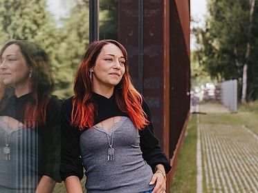 Portraitbild der Germanistik-Studentin Jasmin Lanz, die in grüner Umgebung an eine spiegelnde Gebäudefensterfläche gelehnt ist