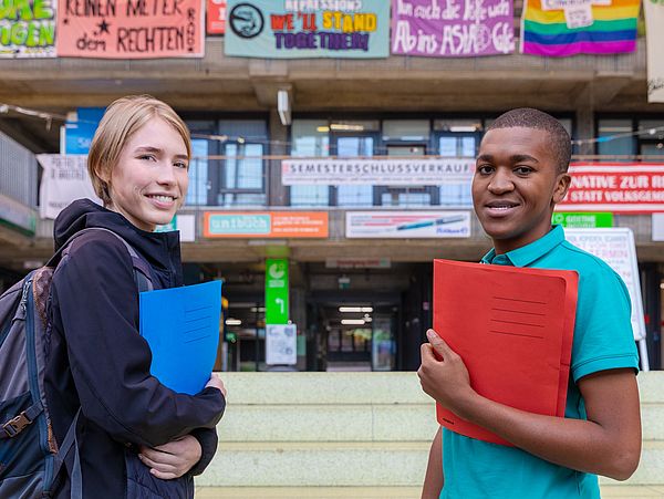 Studierende stehen mit Mappen unterm Arm in der Glashalle und schauen in die Kamera