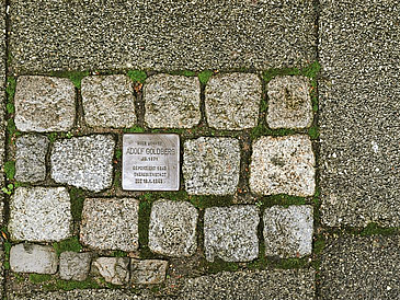 Kopfsteinpflaster auf Bürgerstein, ein Stein wurde durch gravierte Metallplatte ersetzt ("Stolperstein")