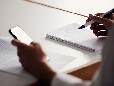Eine Person hält in der linken Hand ein Handy, in der rechten Hand einen Stift, um etwas in das Heft zu schreiben, was ebenfalls zu sehen ist.