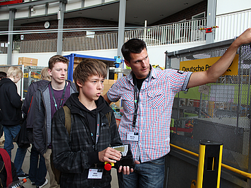 Mann mit mehreren Schülern in Produktionshalle, ein Schüler hält Steuerung für Roboter in der Hand.