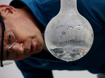 Nahaufnahme von Mann mit Brille, der auf einen Glaskolben schaut, in dem kondensiertes Wasser zu sehen ist.