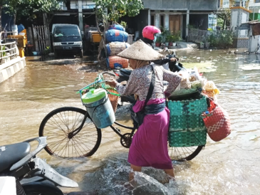Eine Frau schiebt ein mit bunten geflochteten Körben und Taschen schwer bepacktes Fahrrad durch eine von Wasser überschwemmte Straße